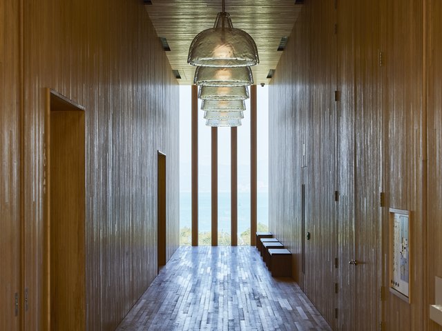 A corridor with a wooden floor and wooden walls. Glass lamps hang from the ceiling in a row. Benches line the wall next to a floor-to-ceiling window with four wooden grilles.